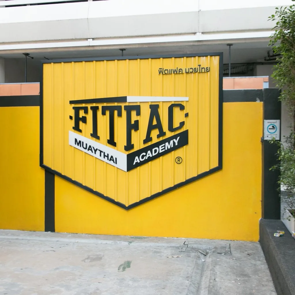 โลโก้โล่ FITFAC Muaythai Academy บนผนังทางเข้าสีเหลือง — FITFAC สาขาเพลินจิต